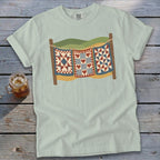 Light green t-shirt featuring a graphic of three patchwork quilts hanging on a clothesline outdoors.