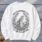 A white crewneck sweatshirt featuring a black graphic of a bird on a fence post, surrounded by corn stalks and the text WHEN THE WHIPPOORWILL CALLS IT'S TIME TO PLANT CORN.