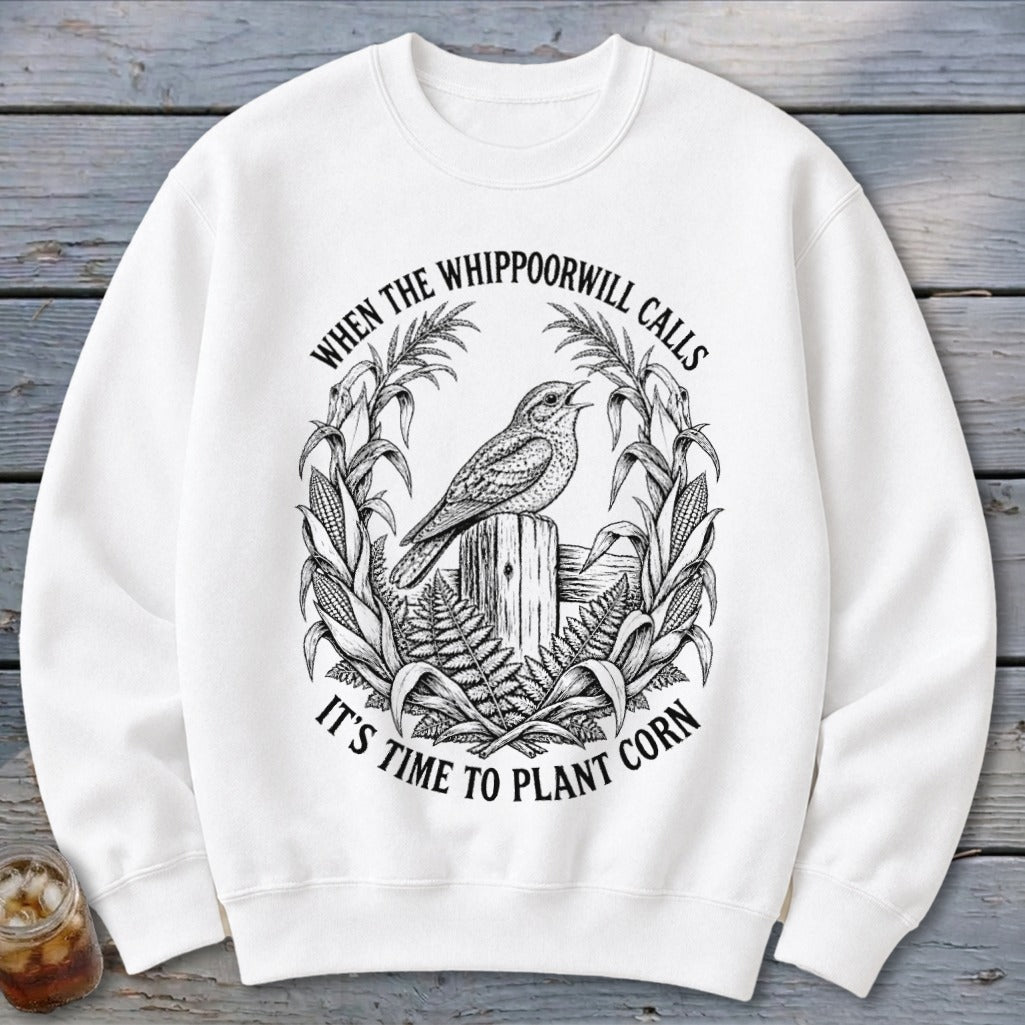 A white crewneck sweatshirt featuring a black graphic of a bird on a fence post, surrounded by corn stalks and the text WHEN THE WHIPPOORWILL CALLS IT'S TIME TO PLANT CORN.