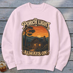 Pink sweatshirt with a graphic of a cabin at sunset and text "The Porch Light is Always On".