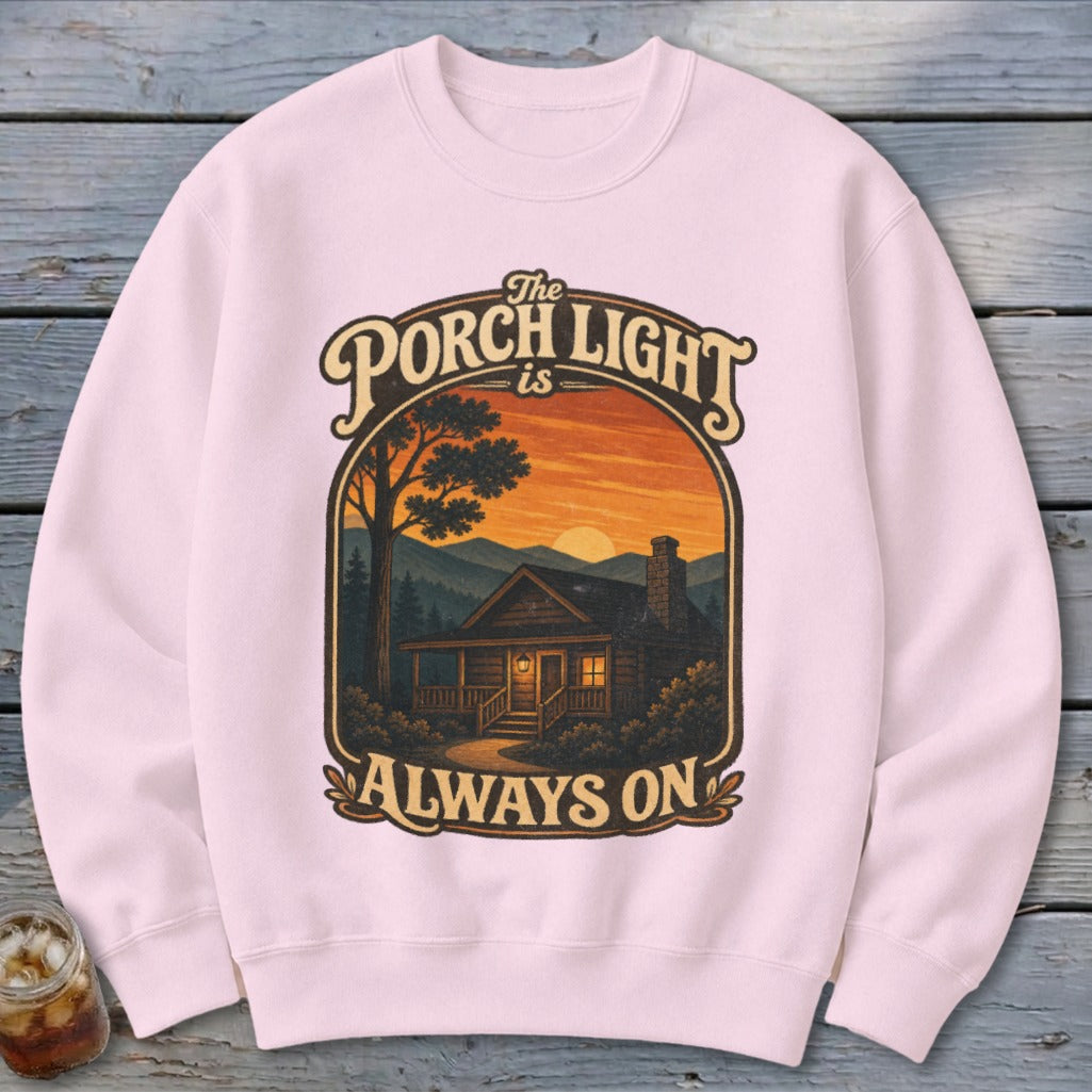 Pink sweatshirt with a graphic of a cabin at sunset and text "The Porch Light is Always On".