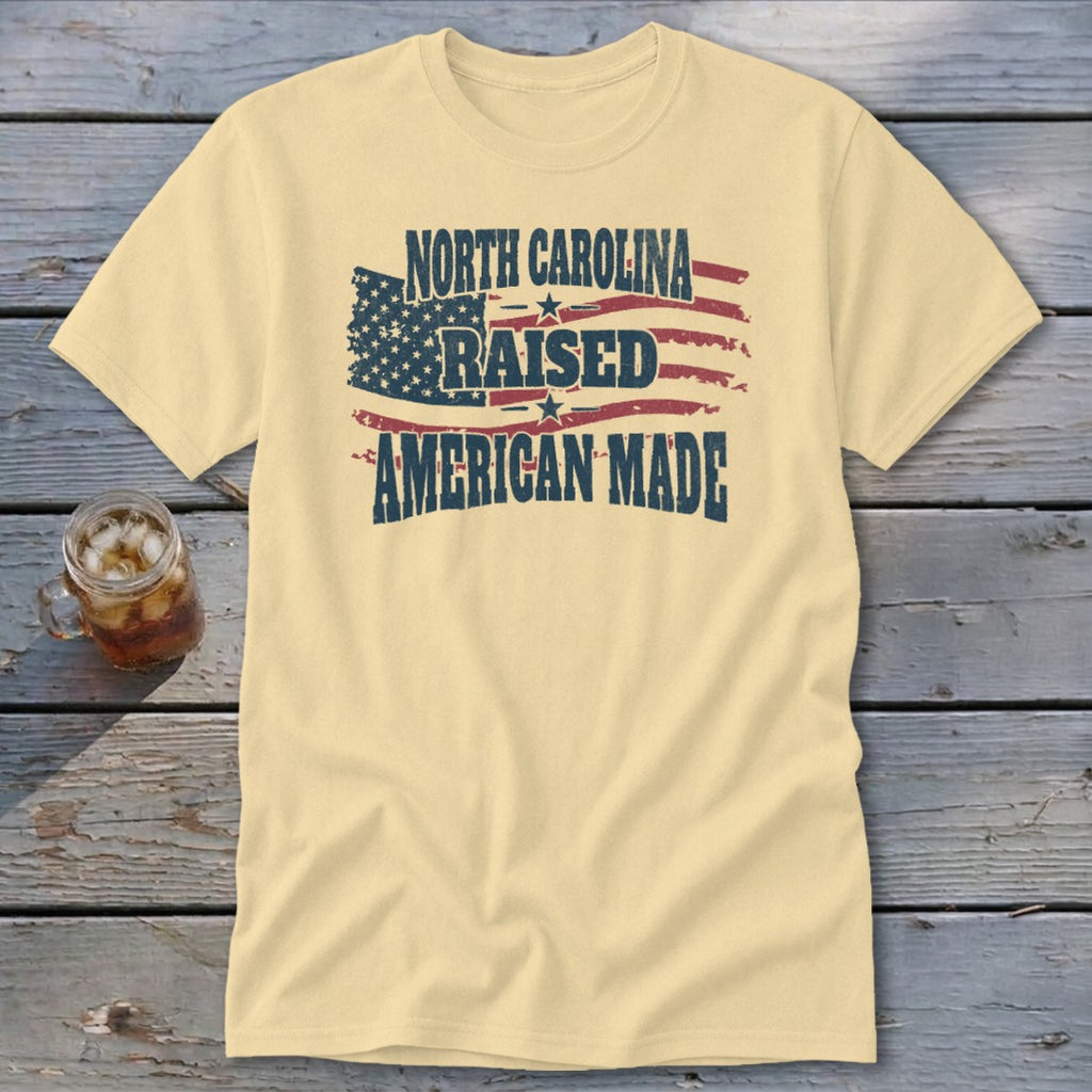 A pale yellow t-shirt features a distressed graphic of an American flag with text reading North Carolina Raised American Made.