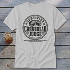 A light gray t-shirt with a round black graphic reading "Certified Cornbread Judge" on a wooden surface.