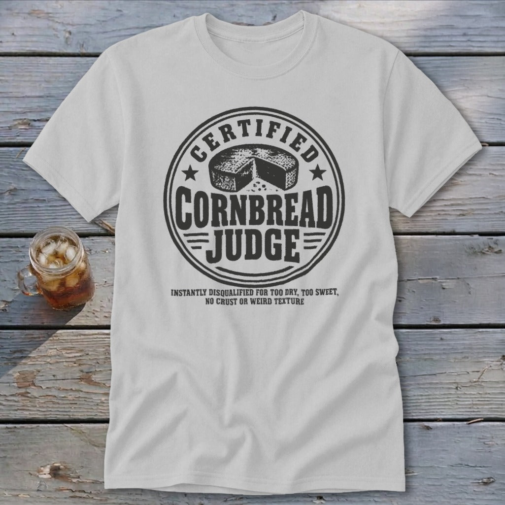 A light gray t-shirt with a round black graphic reading "Certified Cornbread Judge" on a wooden surface.