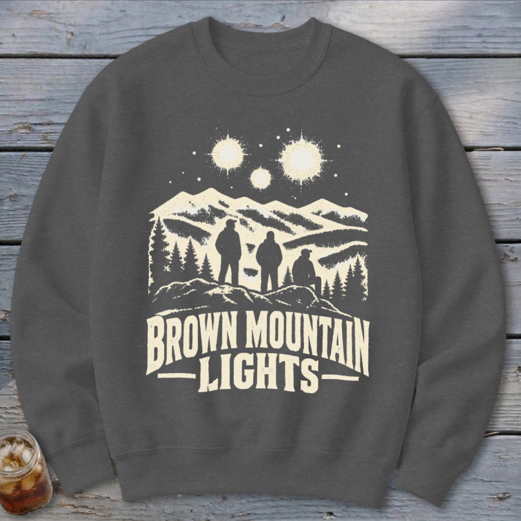 Dark grey crewneck sweatshirt featuring a graphic of three figures, mountains, and three glowing lights with the text Brown Mountain Lights.