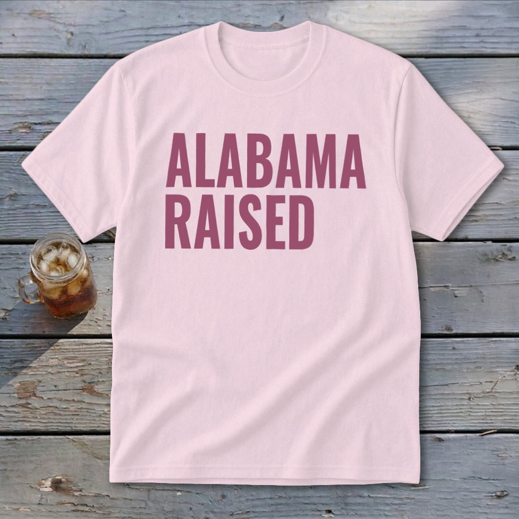 Pink t-shirt with maroon text reading "Alabama Raised" next to a glass of iced tea.