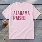 Pink t-shirt with maroon text reading "Alabama Raised" next to a glass of iced tea.