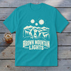 Teal t-shirt featuring a white graphic of three figures, mountains, glowing orbs, and text reading BROWN MOUNTAIN LIGHTS.