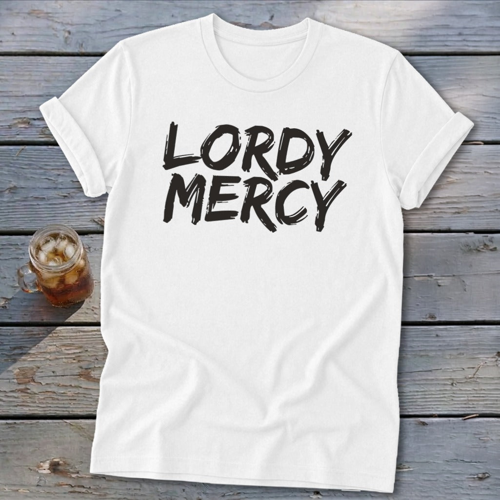 White t-shirt with "LORDY MERCY" text in black, placed on wooden planks with a glass of iced tea.