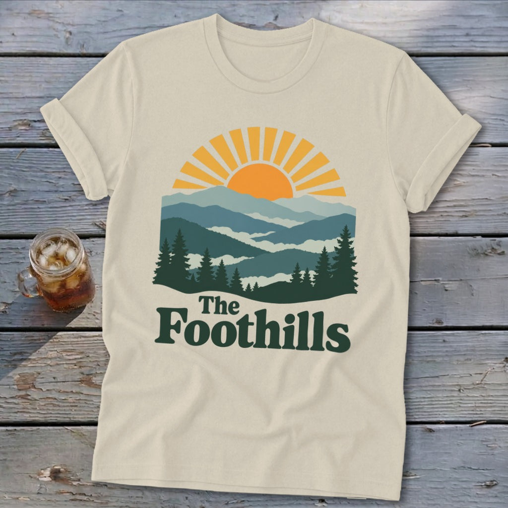 Cream t-shirt with graphic of mountains, sun, and text "The Foothills".