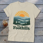 Cream t-shirt with graphic of mountains, sun, and text "The Foothills".