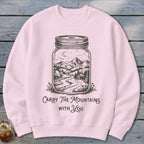 A light pink crewneck sweatshirt featuring a black graphic of a mountain landscape inside a jar with the text Carry The Mountains With You.