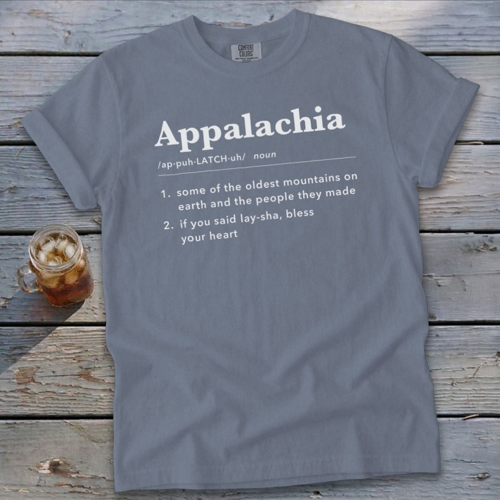 A gray crewneck t-shirt featuring dictionary-style text defining Appalachia, laid flat on a rustic wooden surface with a glass of iced tea.