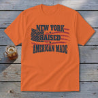 Orange crew neck t-shirt with distressed text reading New York Raised American Made over a US flag graphic.