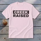 Light pink t-shirt with black text "CREEK RAISED" on a dark rectangle.