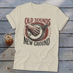 A light-colored t-shirt with a retro graphic of a hand playing a banjo and text reading Old Sounds New Ground.