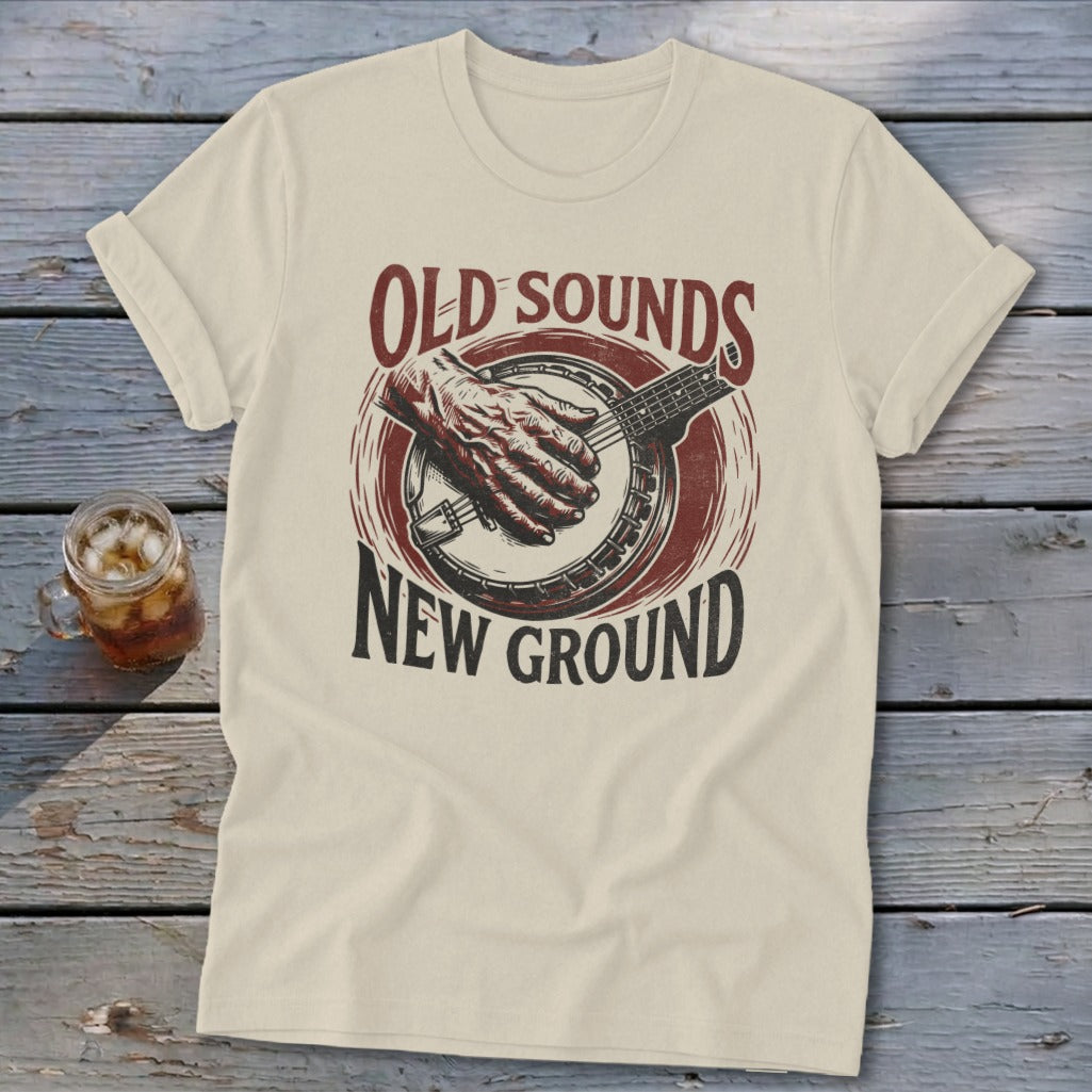 A light-colored t-shirt with a retro graphic of a hand playing a banjo and text reading Old Sounds New Ground.