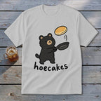 A light gray t-shirt featuring a cartoon bear flipping a pancake with the text hoecakes.