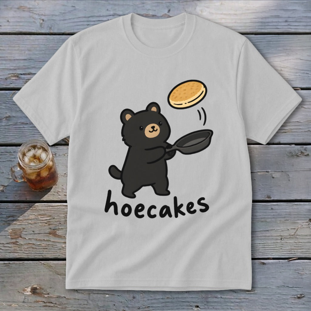 A light gray t-shirt featuring a cartoon bear flipping a pancake with the text hoecakes.