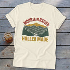 Cream-colored t-shirt featuring a retro mountain landscape graphic with the text Mountain Raised Holler Made.