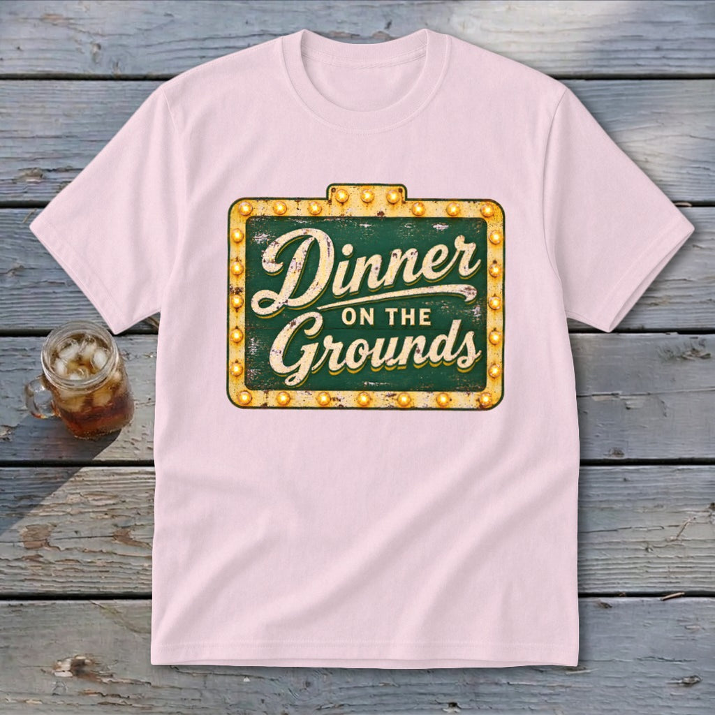 Light pink short-sleeved t-shirt featuring a green vintage sign graphic with lit lightbulbs and text reading Dinner on the Grounds.