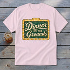 Light pink short-sleeved t-shirt featuring a green vintage sign graphic with lit lightbulbs and text reading Dinner on the Grounds.