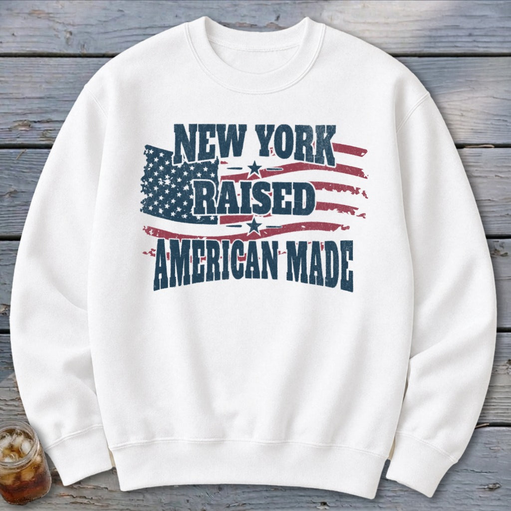 A white crewneck sweatshirt featuring a distressed American flag graphic with the text "New York Raised American Made."