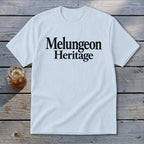 A light blue t-shirt with the black text "Melungeon Heritage" centered on the chest, laid flat on a rustic wooden surface next to a glass of iced tea.