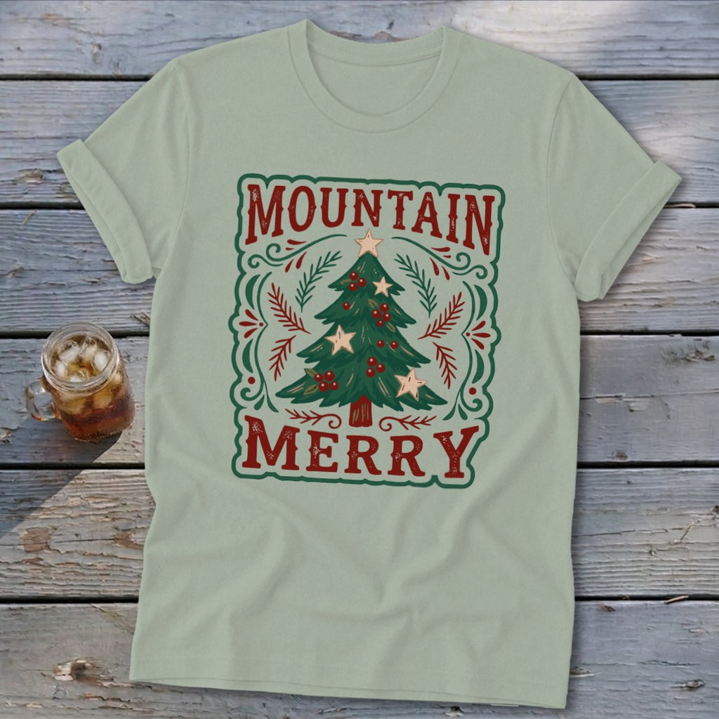 A sage green t-shirt displays a vintage-style graphic with the red text Mountain Merry surrounding a decorated Christmas tree.