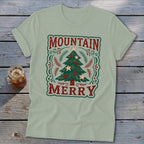 A sage green t-shirt displays a vintage-style graphic with the red text Mountain Merry surrounding a decorated Christmas tree.