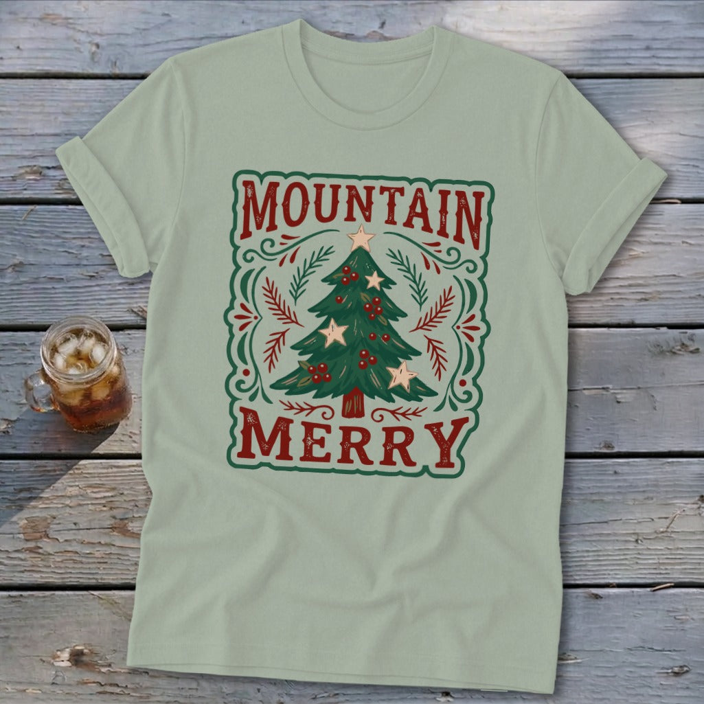 A sage green t-shirt displays a vintage-style graphic with the red text Mountain Merry surrounding a decorated Christmas tree.