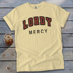 Light yellow t-shirt with "LORDY MERCY" text, on a wooden background with a drink.