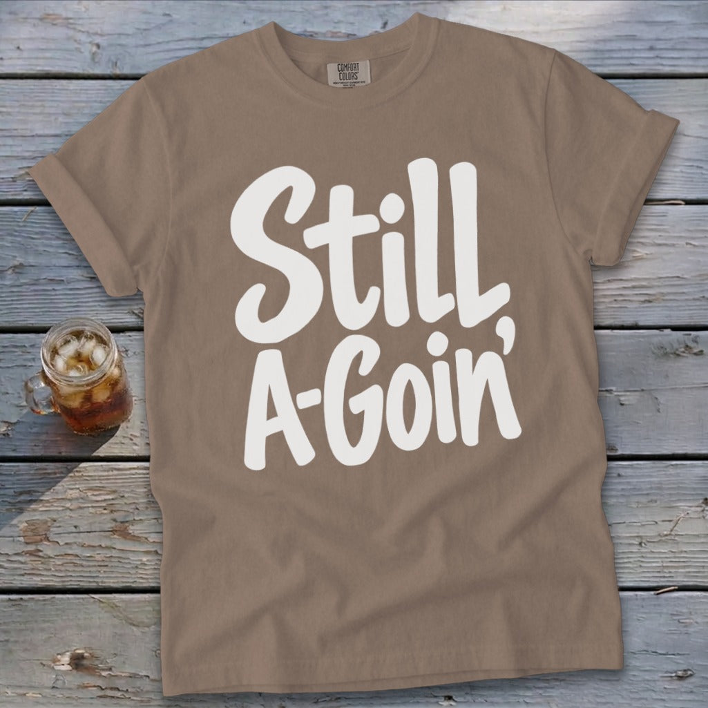 Brown t-shirt featuring the white text "Still A-Goin'" lying flat on a weathered wooden surface.