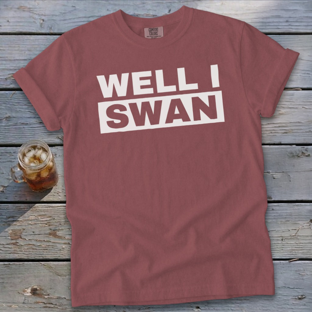 A dusty rose t-shirt featuring the text WELL I SWAN lies flat on a rustic wooden surface next to a glass of iced tea.