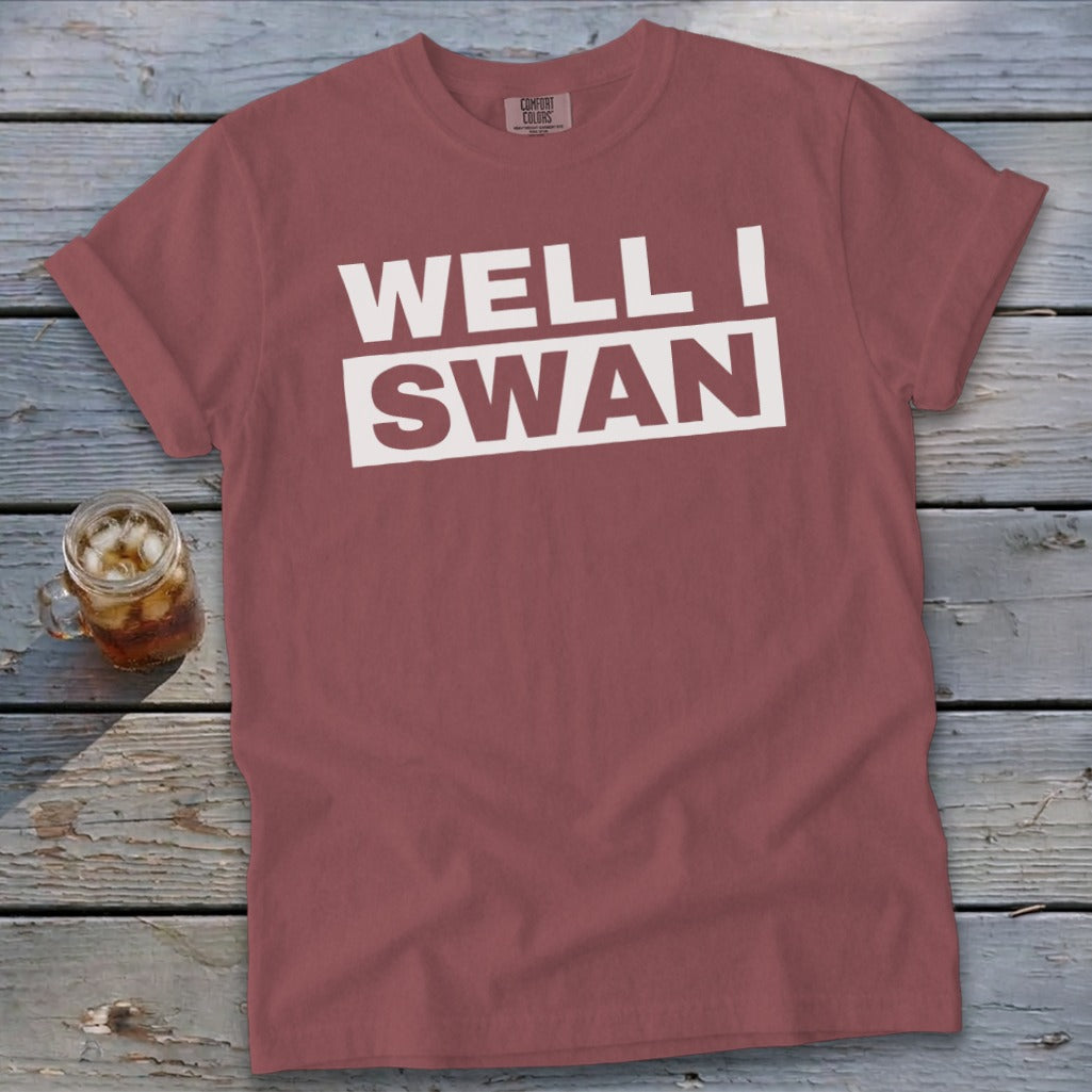 A dusty rose t-shirt featuring the text WELL I SWAN lies flat on a rustic wooden surface next to a glass of iced tea.