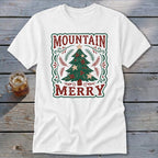 A white t-shirt featuring a green and red holiday graphic with a Christmas tree and text reading MOUNTAIN MERRY.