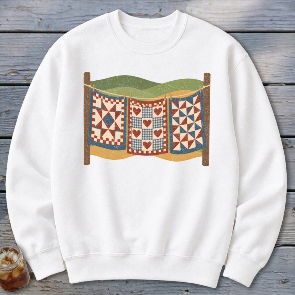 White crewneck sweatshirt with a graphic of three vintage quilts hanging on a clothesline outdoors.
