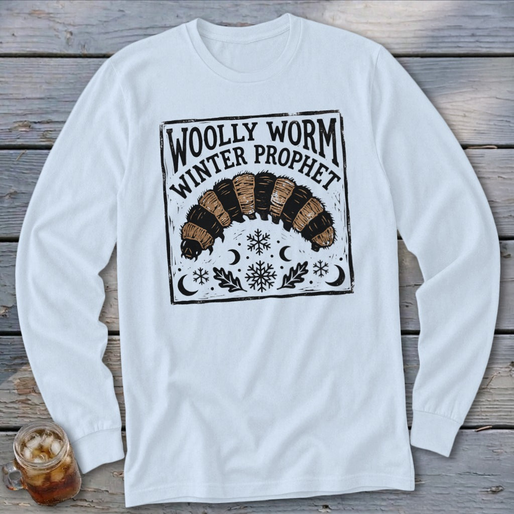 A white long-sleeved t-shirt features a black block-print graphic of a woolly worm with snowflakes, crescent moons, and text reading Woolly Worm Winter Prophet.