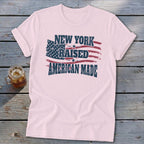 A light pink t-shirt with rolled sleeves features the distressed text New York Raised American Made overlaid on an American flag graphic.