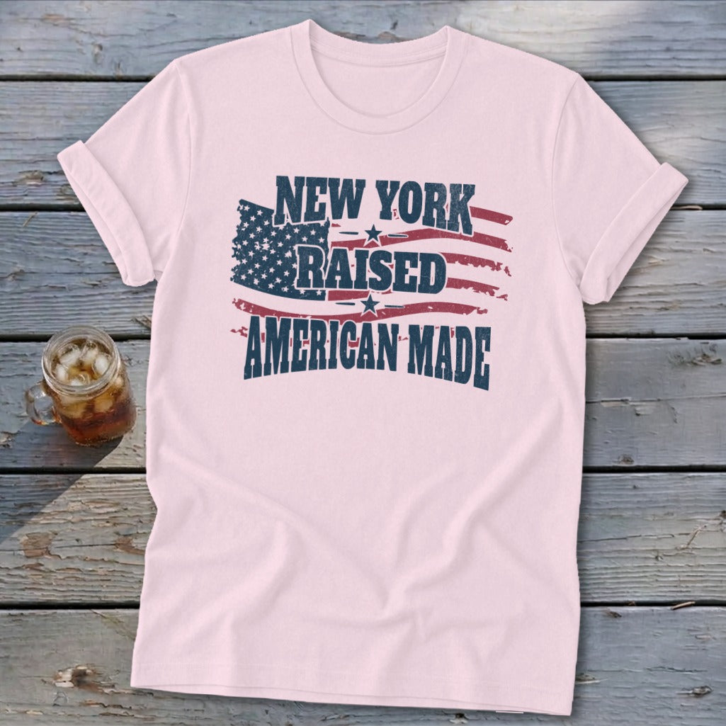 A light pink t-shirt with rolled sleeves features the distressed text New York Raised American Made overlaid on an American flag graphic.