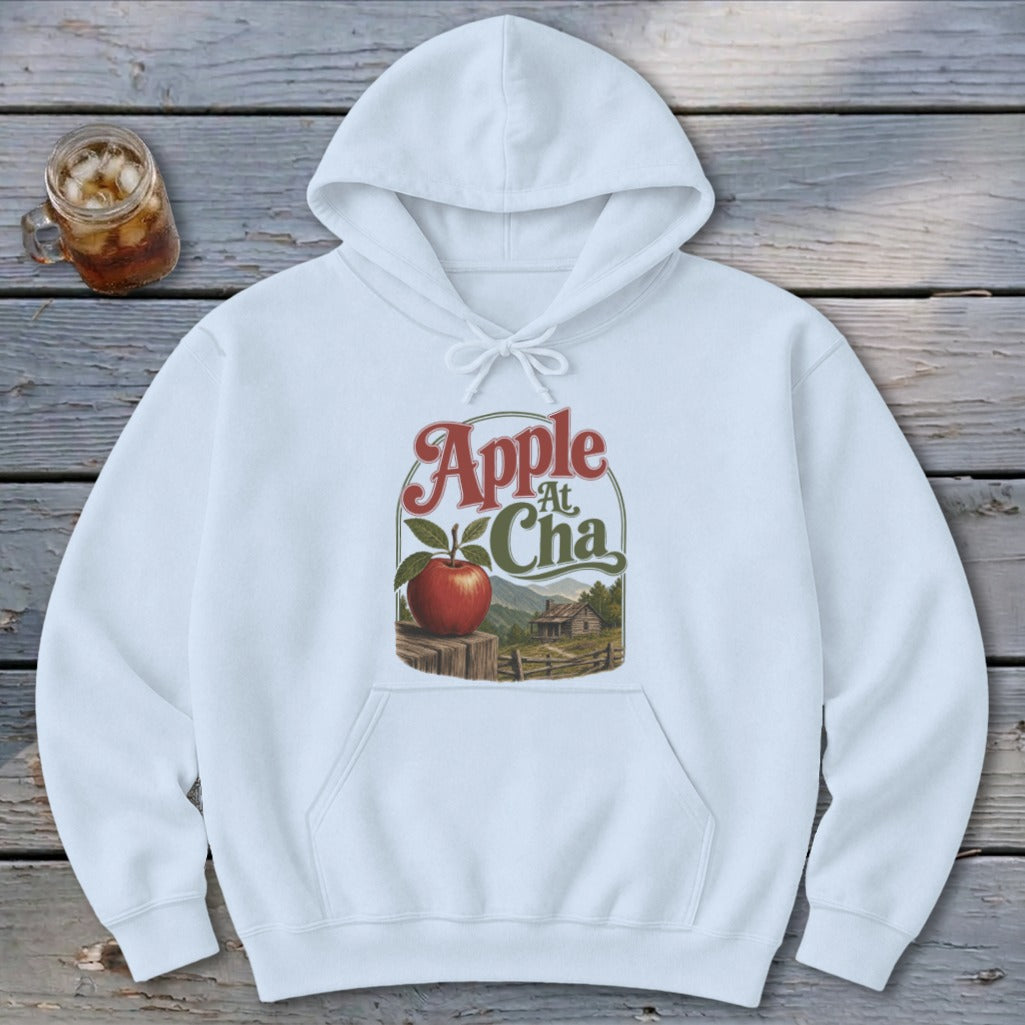 A white hooded sweatshirt with a front pocket, featuring a graphic of an apple, a rustic cabin, and the text Apple At Cha.