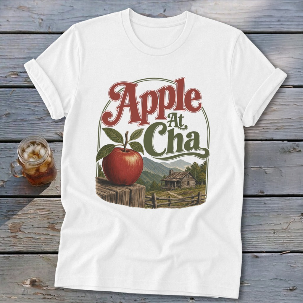 A white t-shirt featuring a graphic with an apple, a cabin, mountains, and the text Apple At Cha.