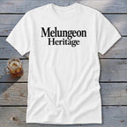 White t-shirt with black text reading Melungeon Heritage, displayed on a weathered wooden surface next to a glass of iced tea.