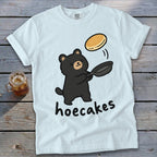 Light blue t-shirt featuring a cartoon black bear flipping a pancake from a skillet, with the text hoecakes underneath.