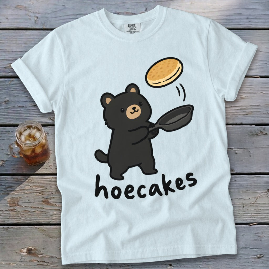 Light blue t-shirt featuring a cartoon black bear flipping a pancake from a skillet, with the text hoecakes underneath.