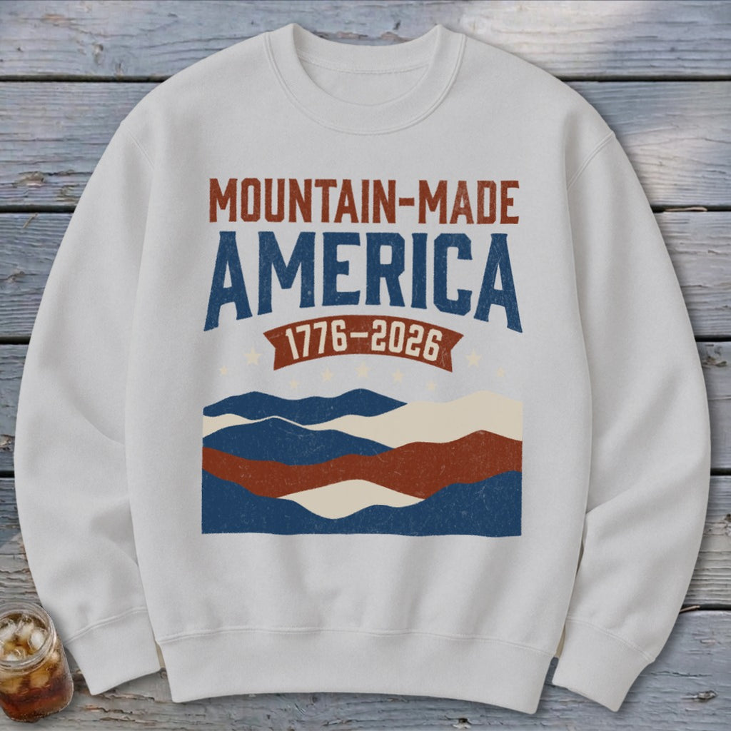 A light gray crewneck sweatshirt featuring a distressed graphic that reads MOUNTAIN-MADE AMERICA 1776-2026 over layered wavy stripes.