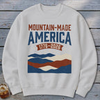 A light gray crewneck sweatshirt featuring a distressed graphic that reads MOUNTAIN-MADE AMERICA 1776-2026 over layered wavy stripes.