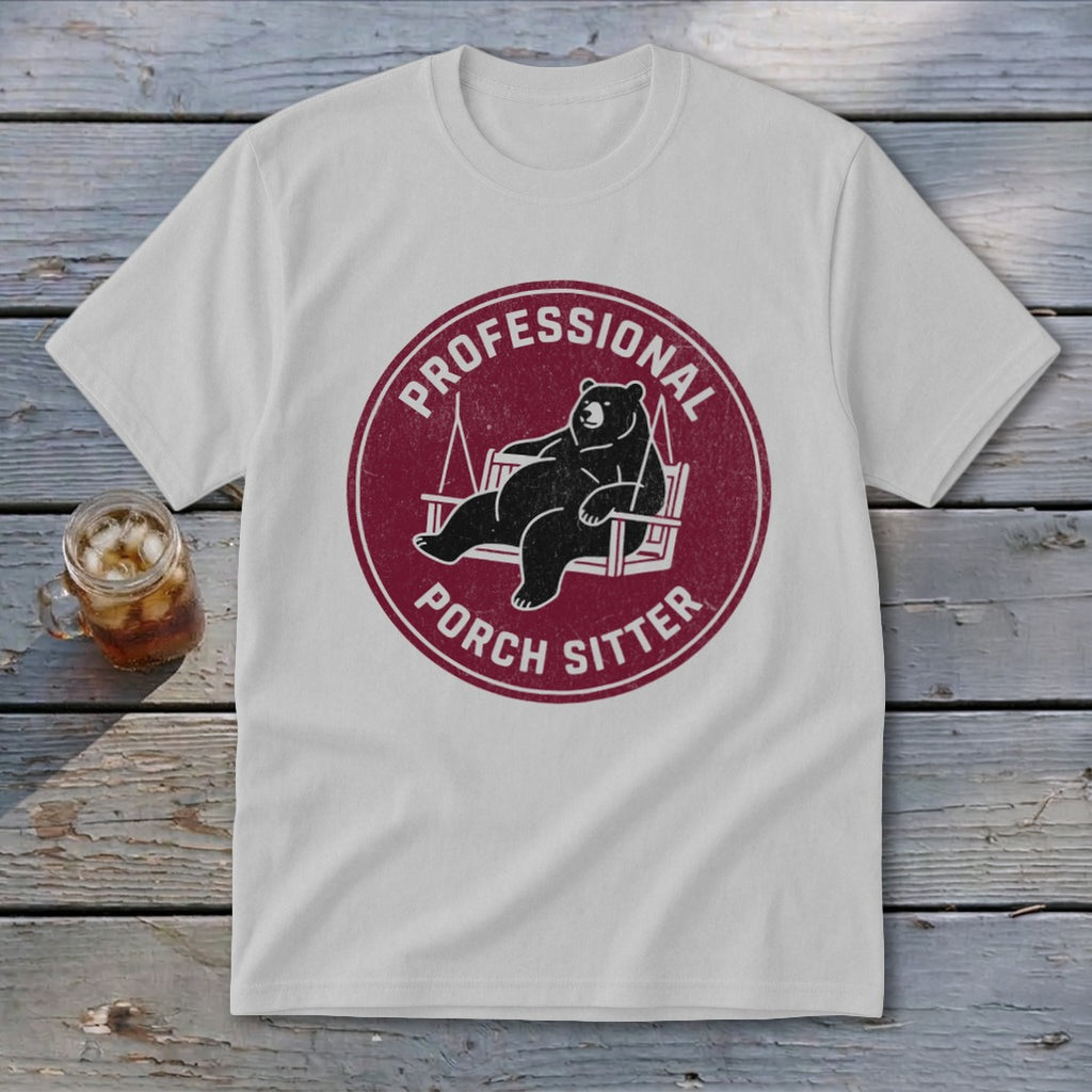 A light gray t-shirt featuring a circular maroon graphic of a bear lounging on a porch swing, accompanied by the text "Professional Porch Sitter".