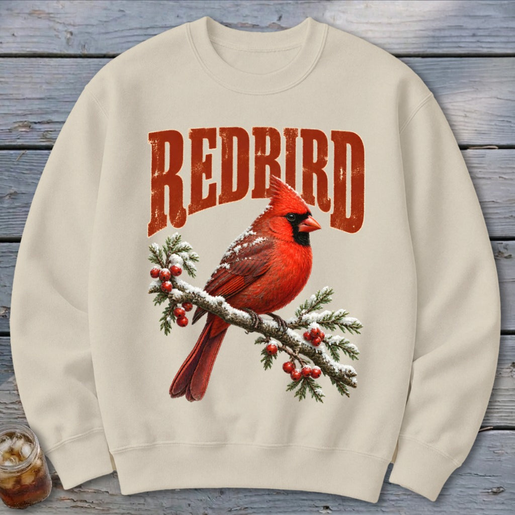 A cream-colored crewneck sweatshirt featuring a graphic of a red cardinal on a snowy branch with red berries and the text REDBIRD above it.