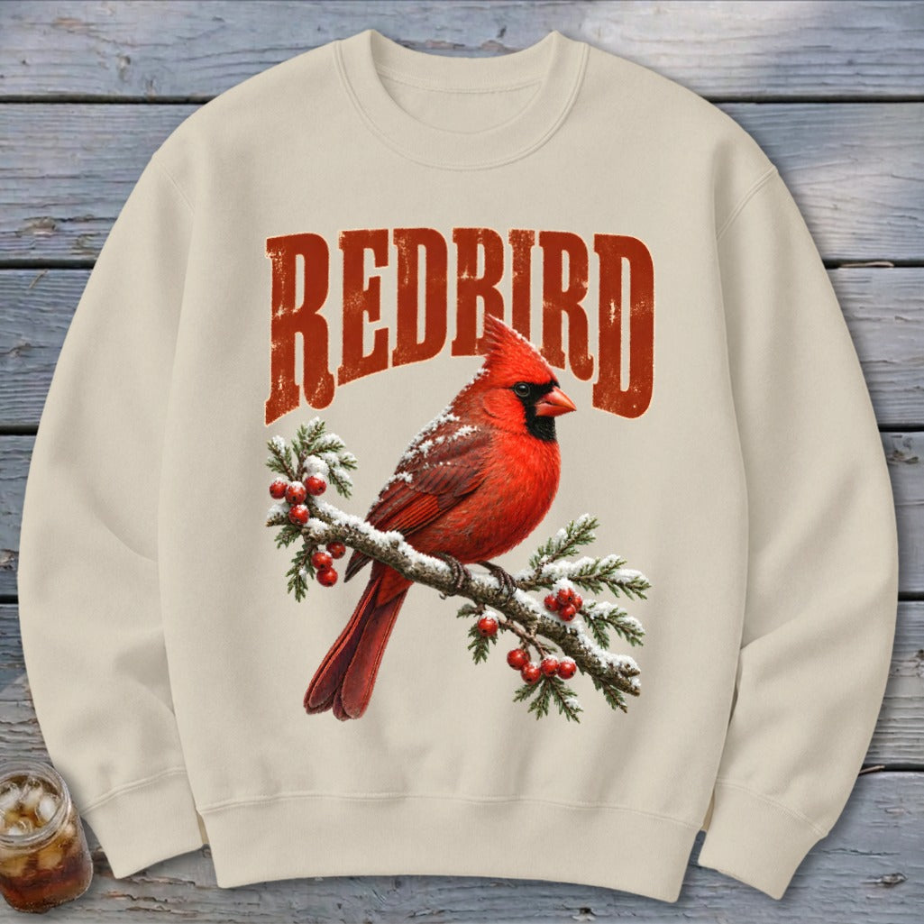 A cream-colored crewneck sweatshirt featuring a graphic of a red cardinal on a snowy branch with red berries and the text REDBIRD above it.